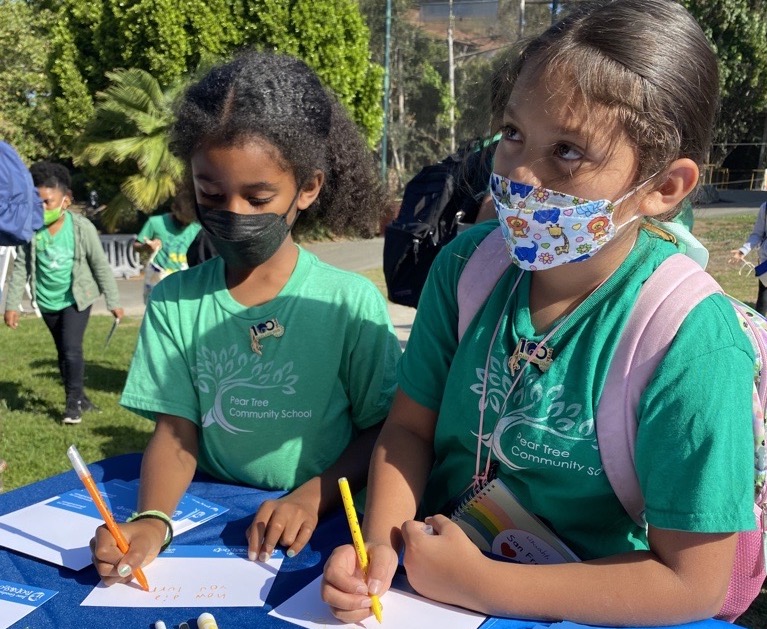 A photo of two children—one with light brown skin and curly, brown hair, the other with tan skin and wavy, brown hair—writing on small pieces of paper in an outdoor space.