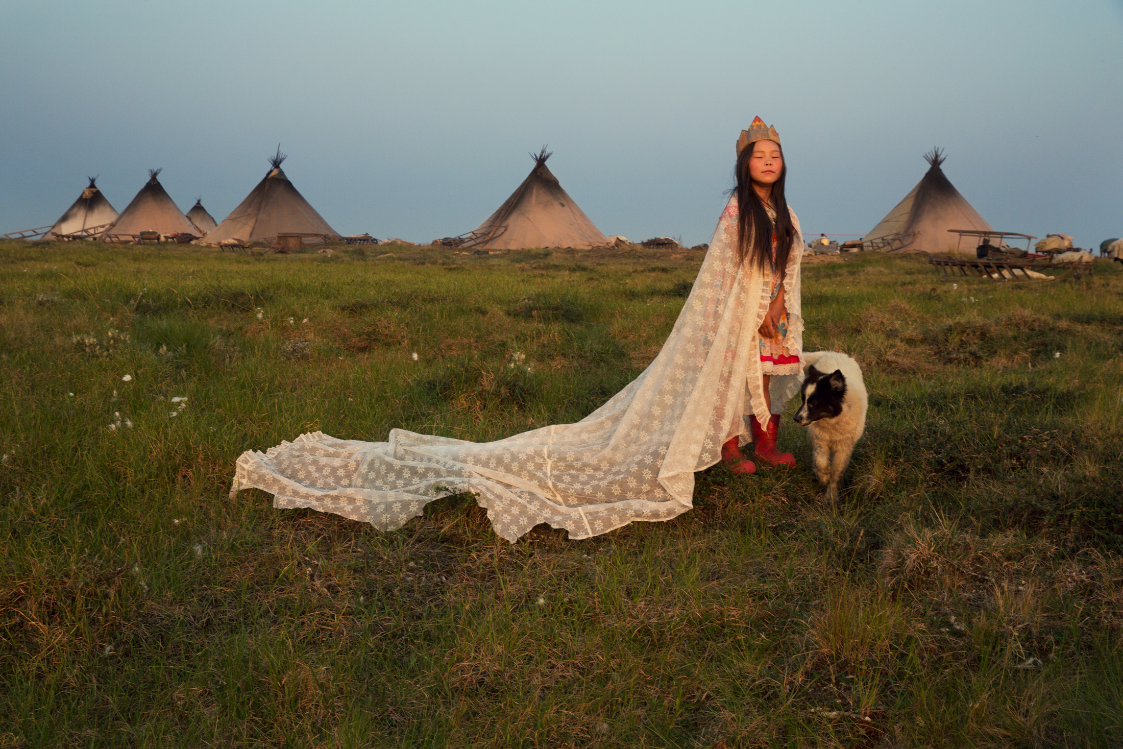 A photo of a young girl with tan skin and straight, black hair in a flowing white cape and paper crown standing next to a dog in a large, open, grassy field. There are large tent structures in the background.