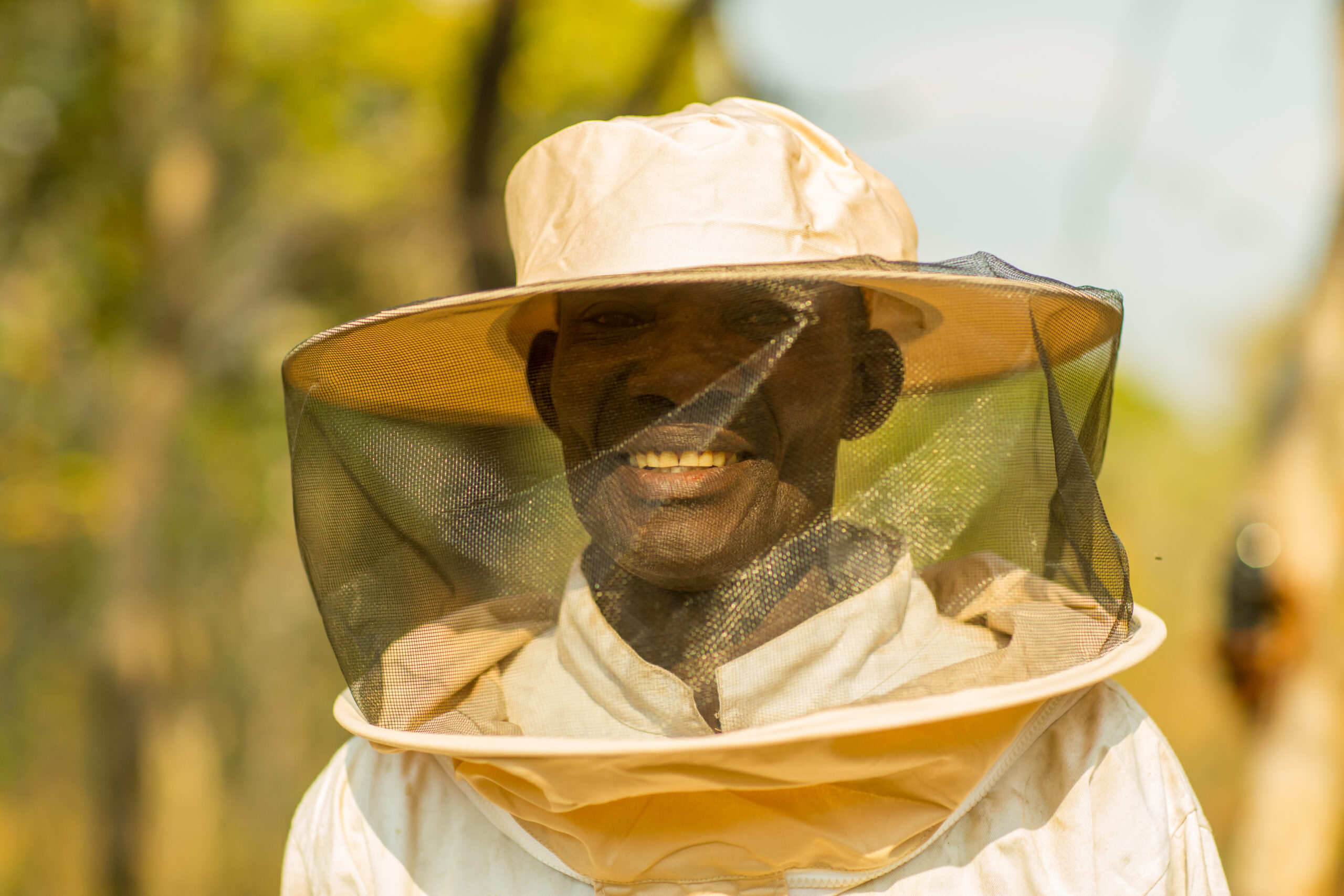 A portrait photo of a middle-aged, masculine person with dark skin, smiling at the camera and wearing a beekeeper outfit that includes a wide-brimmed hat with mesh netting hanging from it.