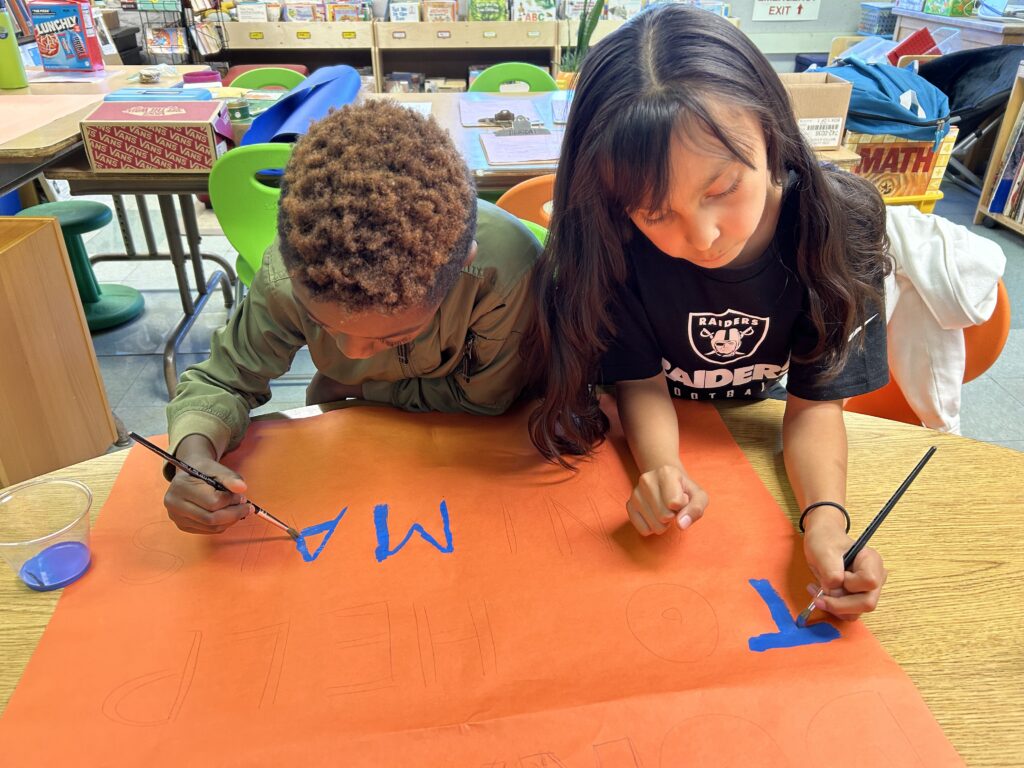 A photo of a child with light brown skin and long, wavy, black hair and a child with dark skin and short, curly, brown hair sitting at a table in a classroom and painting letters on a large poster. 