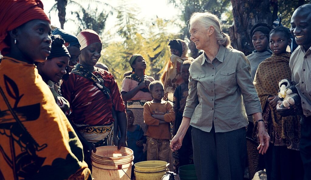A photo of an elderly, feminine person with light skin and white hair in a ponytail (Dr. Jane Goodall) standing outside in the middle of a group of people dressed in colorful clothing.