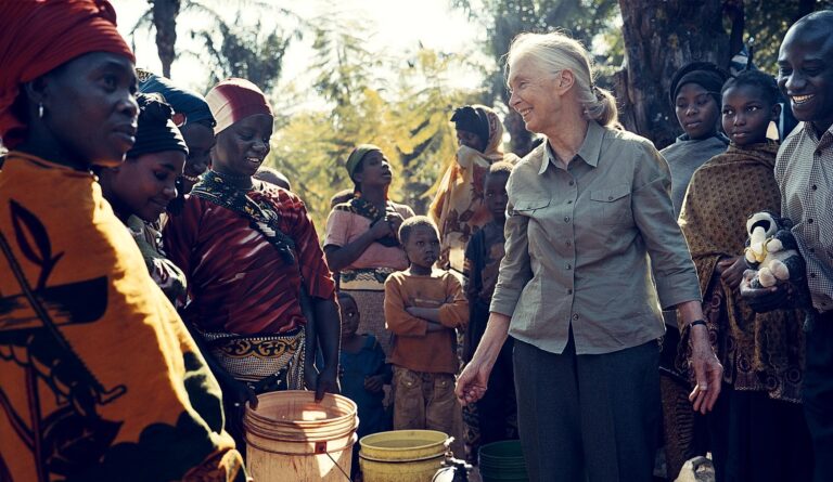 A photo of an elderly, feminine person with light skin and white hair in a ponytail (Dr. Jane Goodall) standing outside in the middle of a group of people dressed in colorful clothing.
