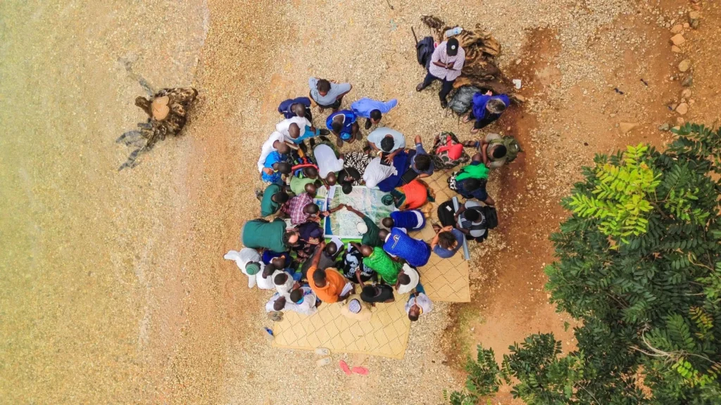 A large group of people in multicolored clothing standing in a circle on a tropical beach pointing at a map.