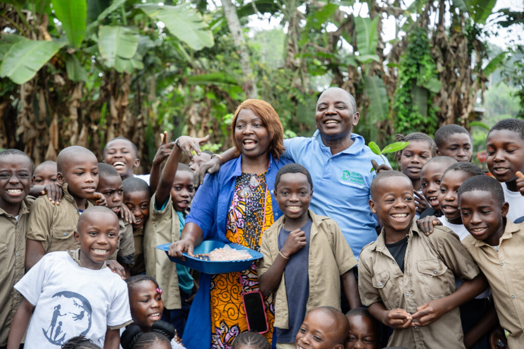 A photo of a middle-aged, feminine person with dark skin and short, red hair standing outside next to a middle-aged, masculine person with dark skin and a shaved head. Both of them are surrounded by a large group of children.