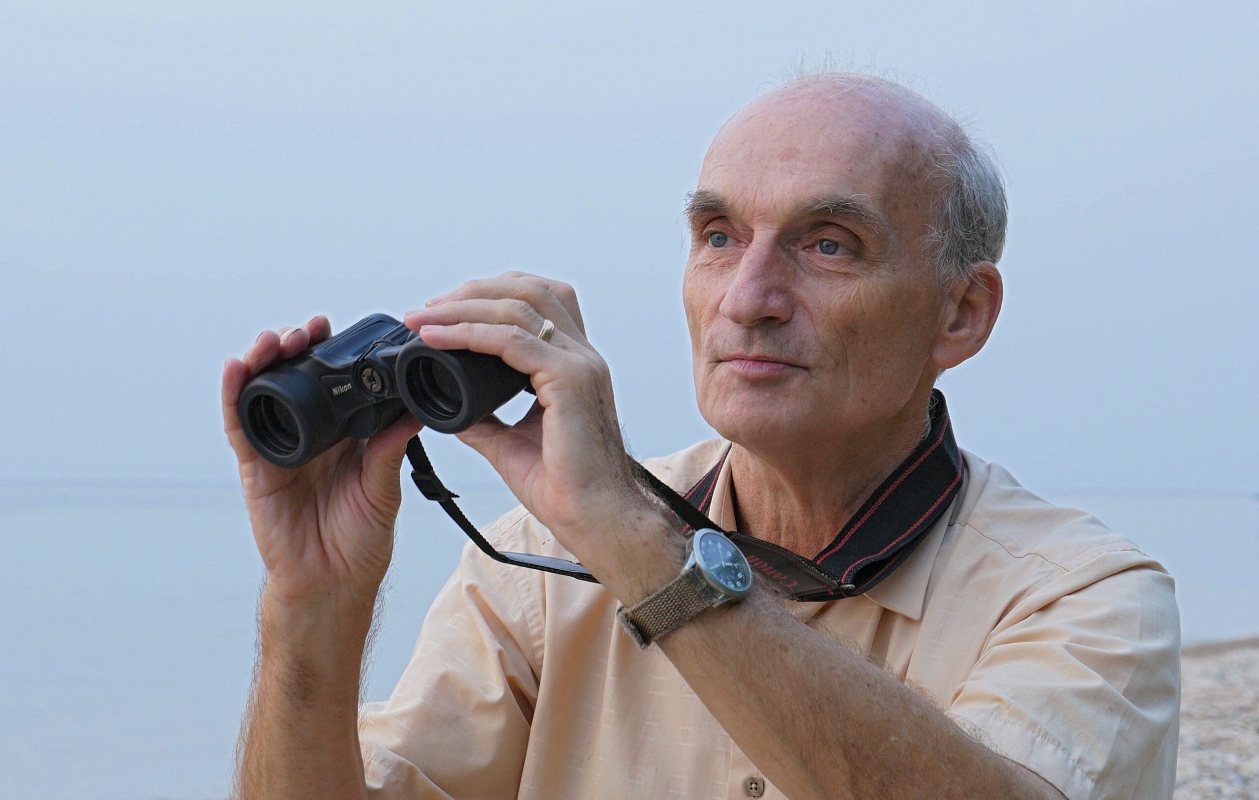 A photo of an elderly, masculine person with light skin and a bald head standing outside and holding up binoculars.