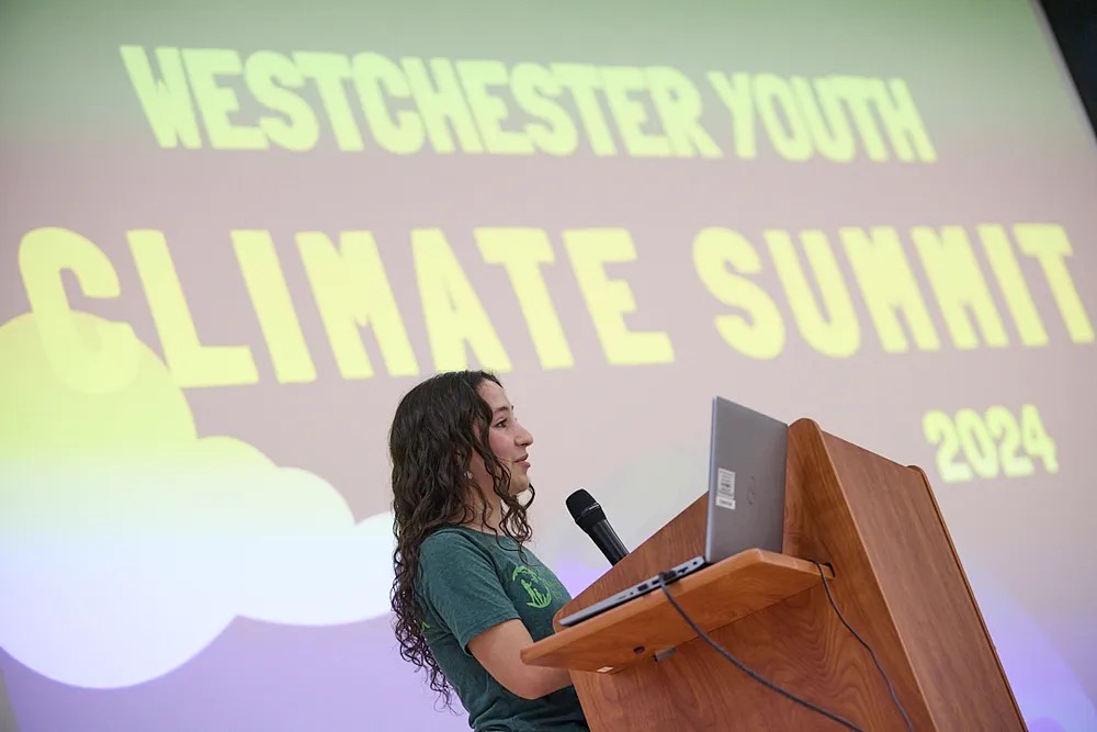 A photo of a teenager with light skin and wavy, brown hair speaking at a podium. On a screen behind her are the words, "Westchester Climate Summit 2024."