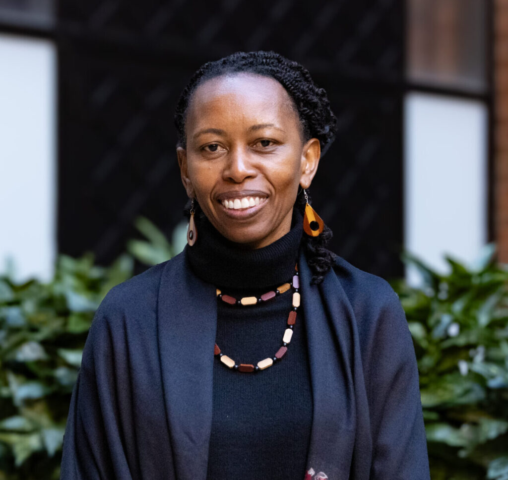 A photo of a middle-aged, feminine person with medium brown skin and black hair pulled back standing outside.