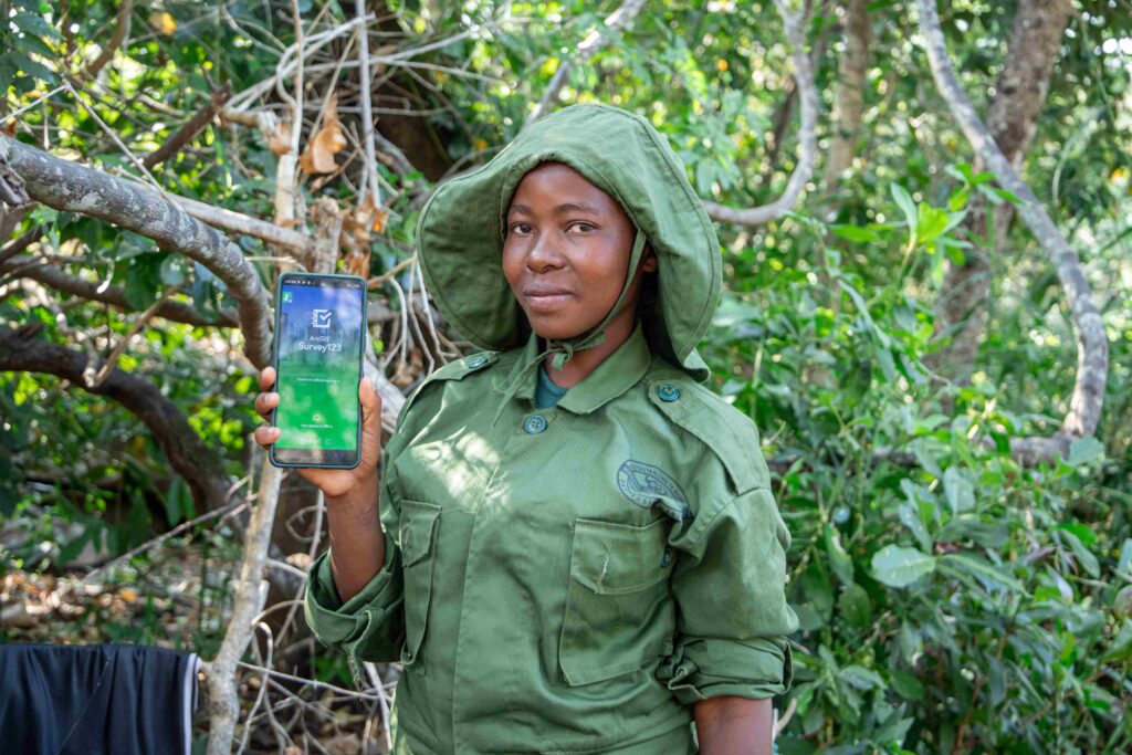 A photo of a young, feminine person with medium brown skin standing in a forest and holding up a smart phone that shows the words, "ArcGIS Survey123."