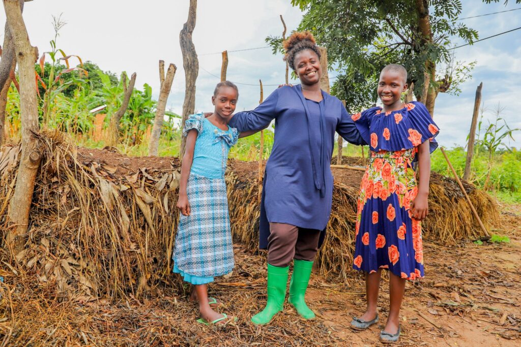 A photo of a young, feminine person with medium brown skin standing outside with two children with medium brown skin in front of a large compost heap.