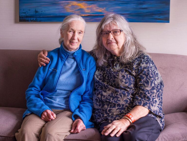 A photo of an elderly, feminine person with shoulder-length, wavy, grey hair sitting on a couch with one arm around an elderly, feminine person with light skin and white hair in a ponytail (Dr. Jane Goodall).