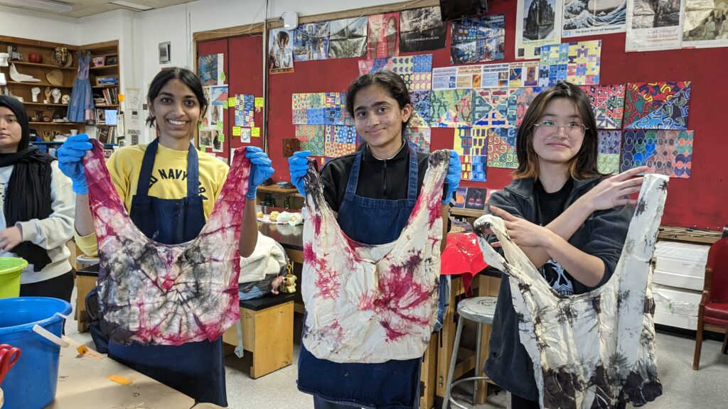 A photo of three teenagers holding up tie-dyed bags in the middle of a room covered in art: a teenager with light brown skin and black hair in a ponytail; a teenager with light brown skin and black hair in a ponytail; a teenager with light skin and shoulder-length, wavy, black hair.