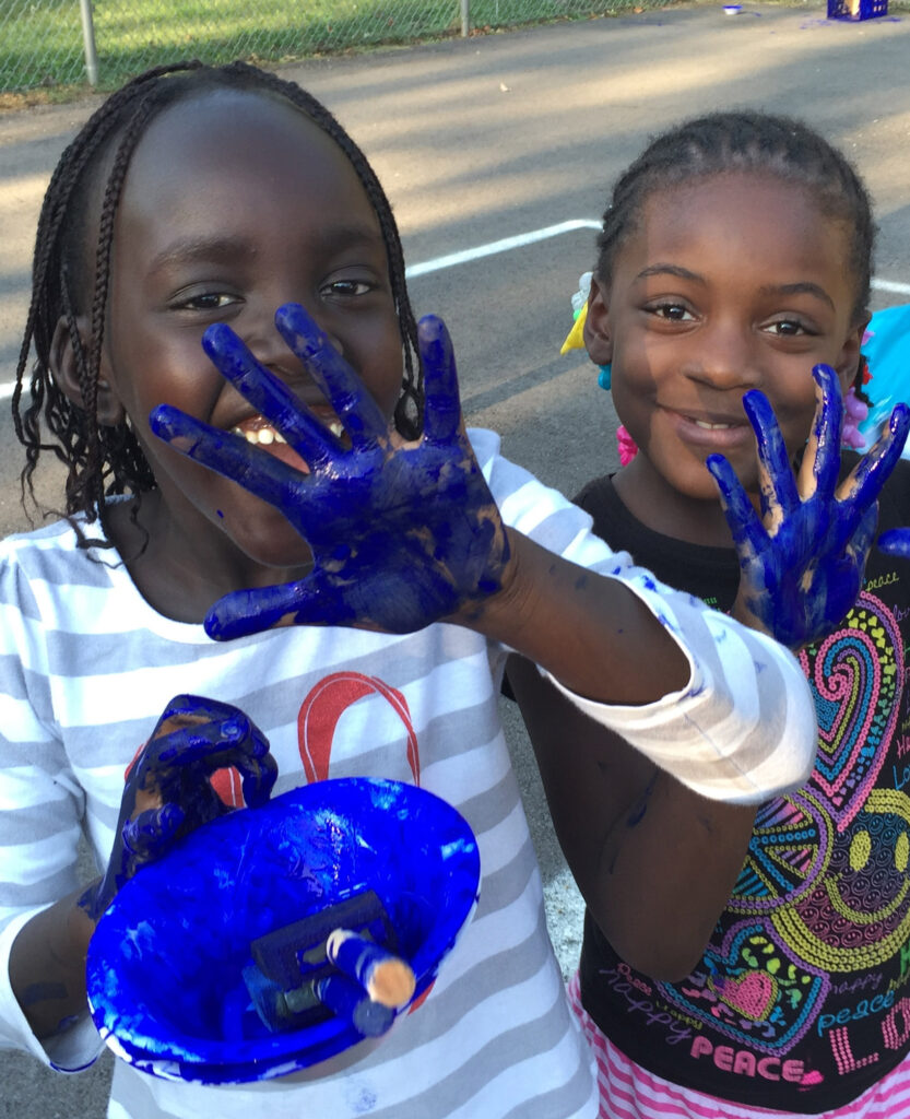 A photo of two children with dark skin and black hair in braids standing outside with blue paint all over their hands.