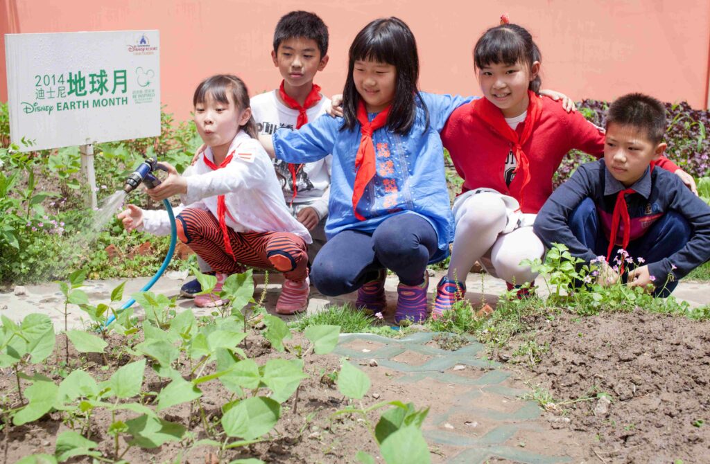 A group of five kids with light skin and straight, black hair sitting outside in a garden and watering the plants.