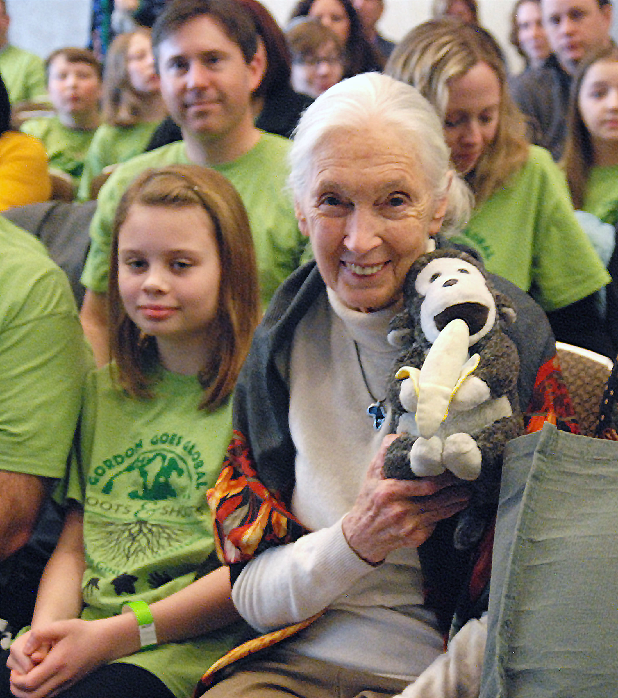 A photo of an elderly, feminine person with light skin and white hair in a ponytail sitting with a group of people in matching green shirts and holding up a plushy monkey toy.