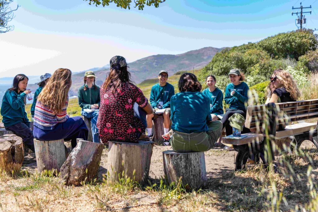 A photo of about ten people of varying appearances sitting on logs outside in a circle and having a conversation.