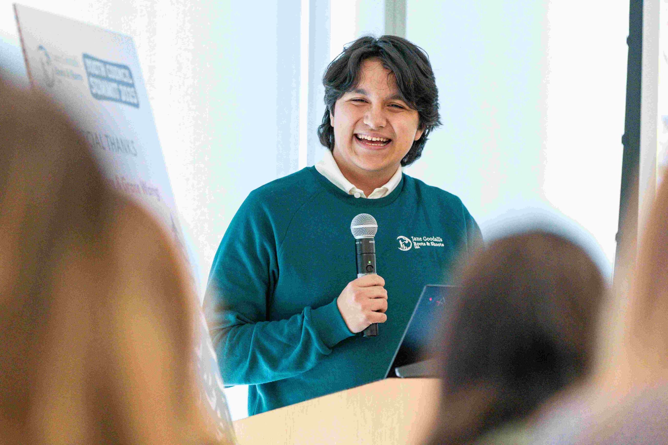 A photo of a teenager with light brown skin and short, wavy, black hair standing at a podium and speaking to a group of people.