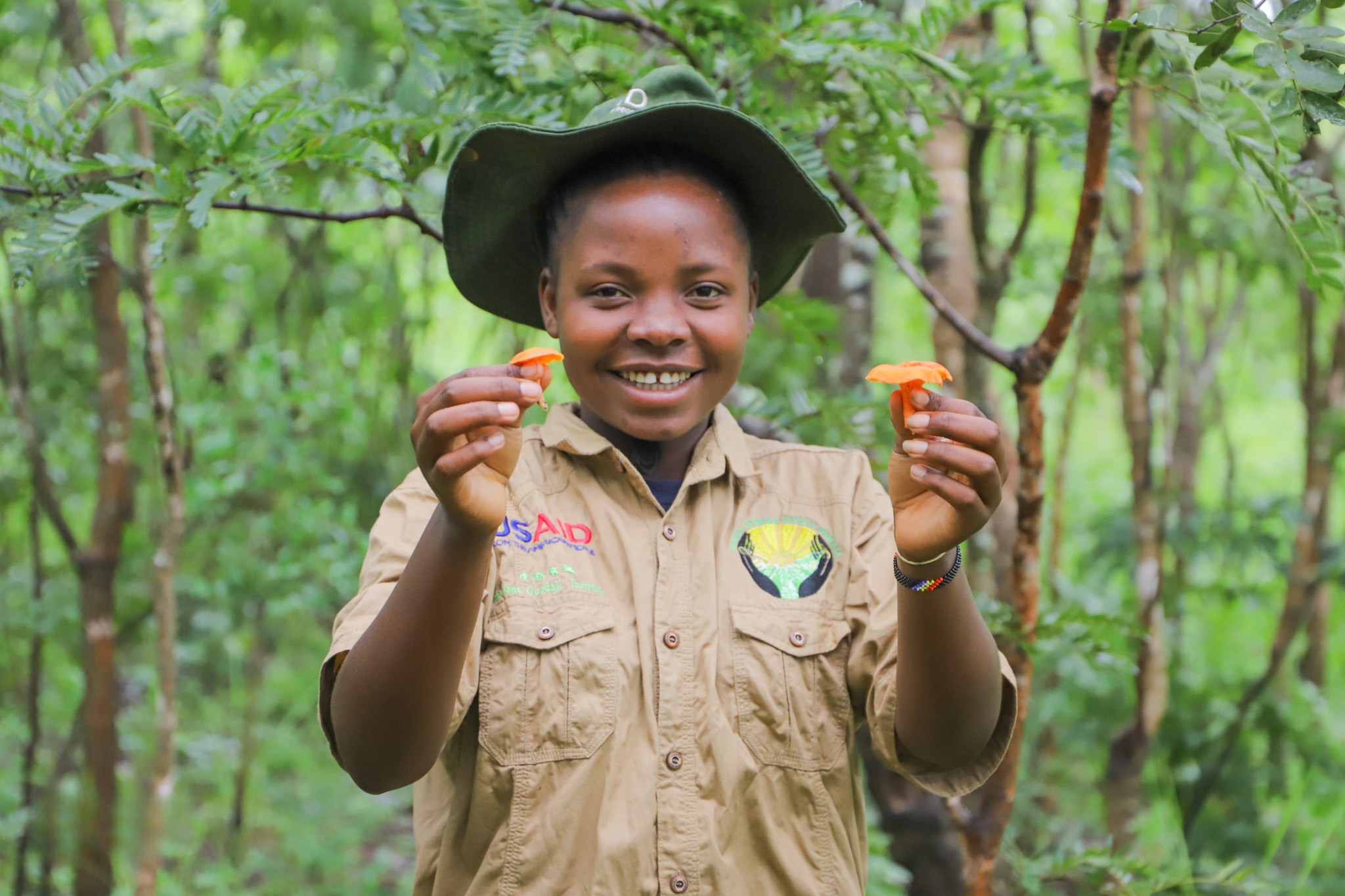 A photo of a young, feminine person with medium brown skin standing in a forest and holding up two bright orange mushrooms.