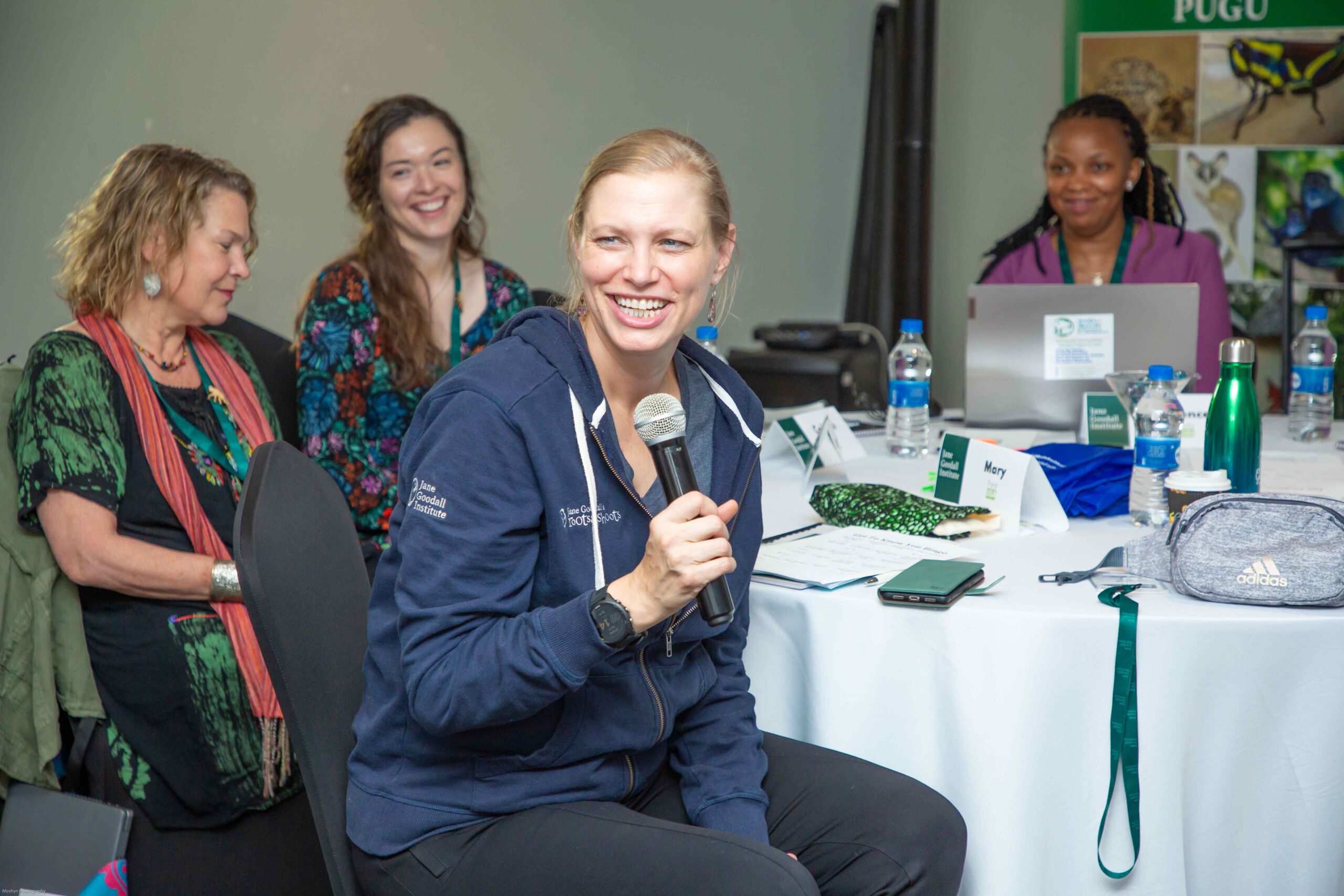 A photo of a middle-aged, feminine person with light skin and blond hair in a ponytail speaking into a microphone at a table of other people.