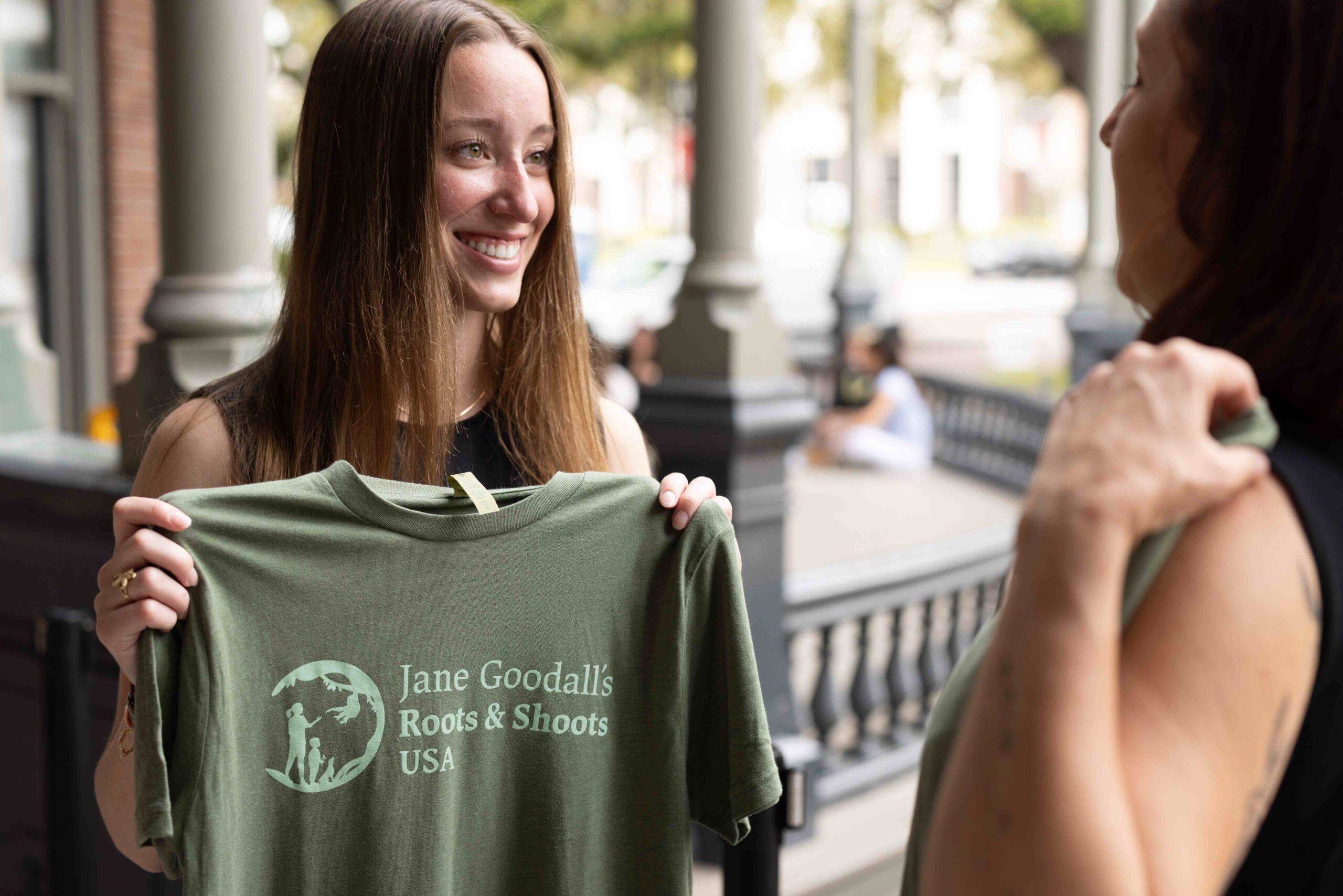 A photo of a young, feminine person with light skin and straight, blond hair standing outside and holding up a shirt that says, "Jane Goodall's Roots & Shoots USA."