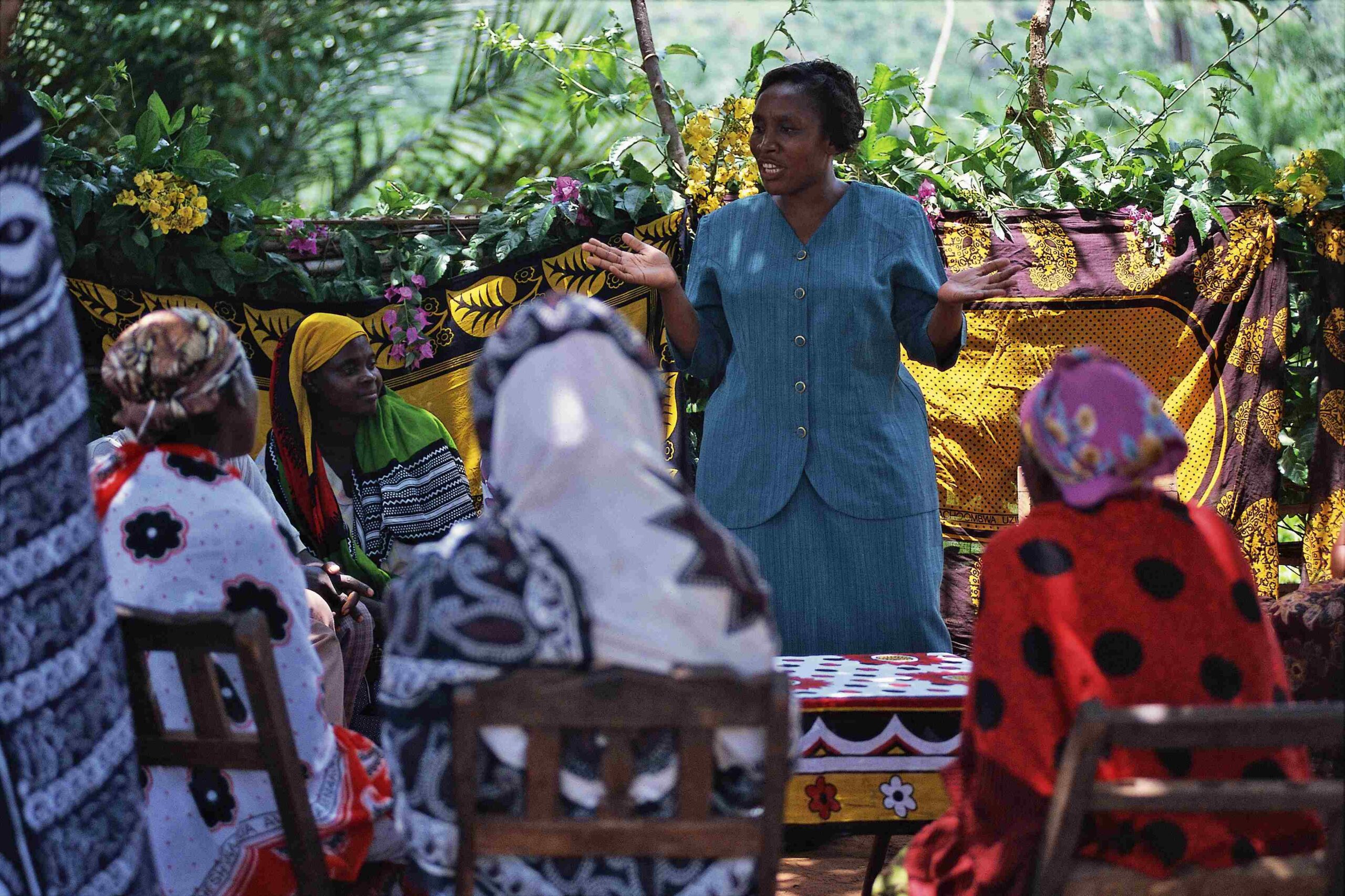 A photo of a middle-aged, feminine person with dark skin and black hair in a bun standing outside and speaking to a group of women.