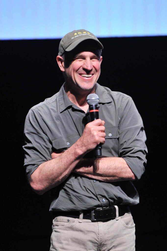 Bill Wallauer, wearing a grey JGI collared shirt while holding a microphone and speaking on stage in front of a large screen.