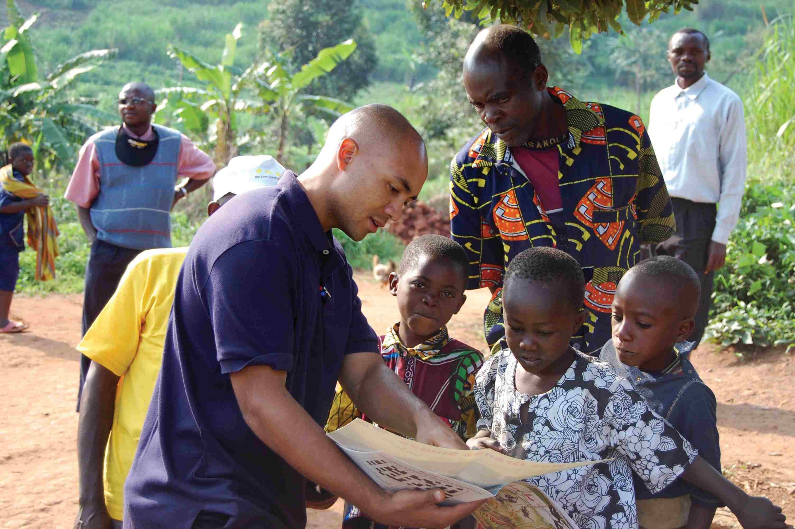 A photo of a young, masculine person with light skin and a shaved head speaking to a group of people outside and gesturing at a map.