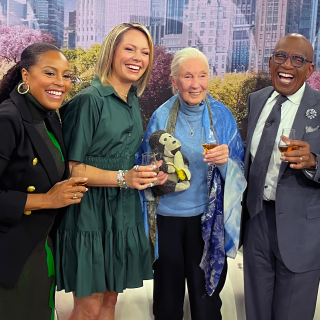 A woman in a blue sweater with a white pony tail stands between three talk show hosts on a news set.
