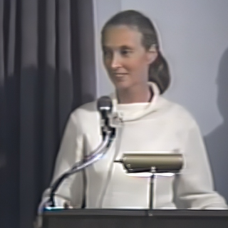 A woman in a white turtleneck with a grey ponytail (Jane Goodall) stands at a podium.