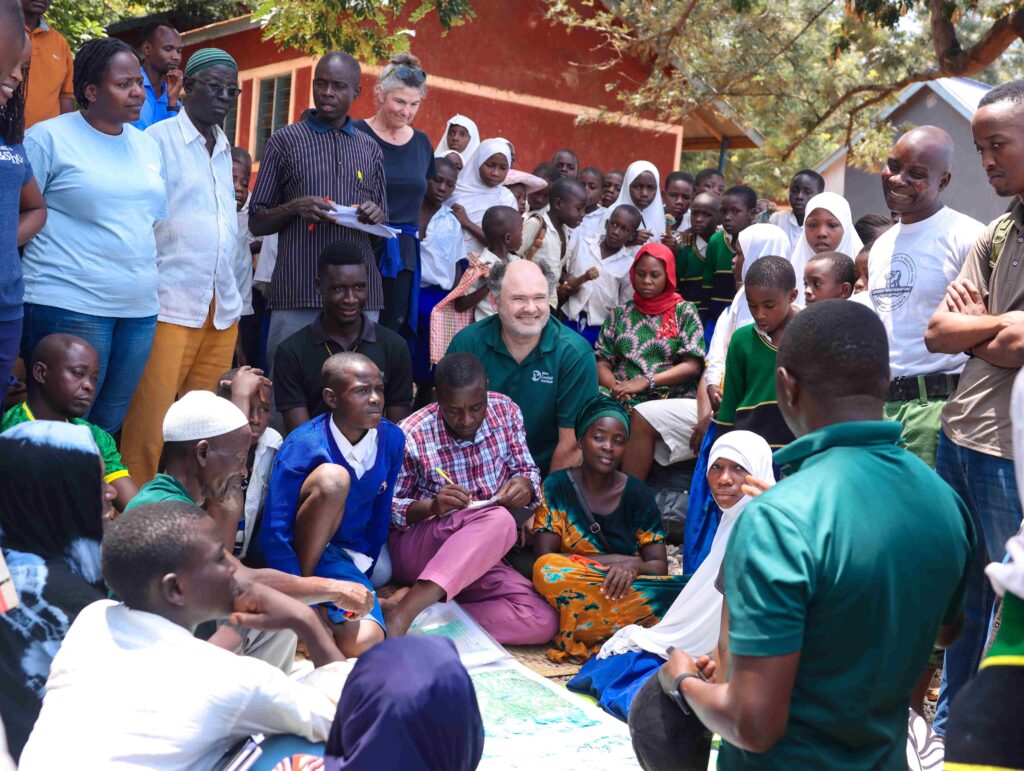 A photo of a large group of people of varying appearances sitting and standing in a circle outside and discussing a map on the ground in the middle of them.