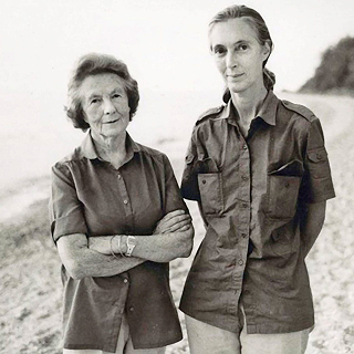A mature woman with short hair (Vanne Goodall) standing next to a younger woman with a pony tail (Jane Goodall) on an isolated beach.