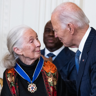 A woman with a white ponytail (Jane Goodall) wearing a medal with a blue ribbon standing next to, and looking into the eyes of a man with white hair and a dark suit (President Joe Biden).