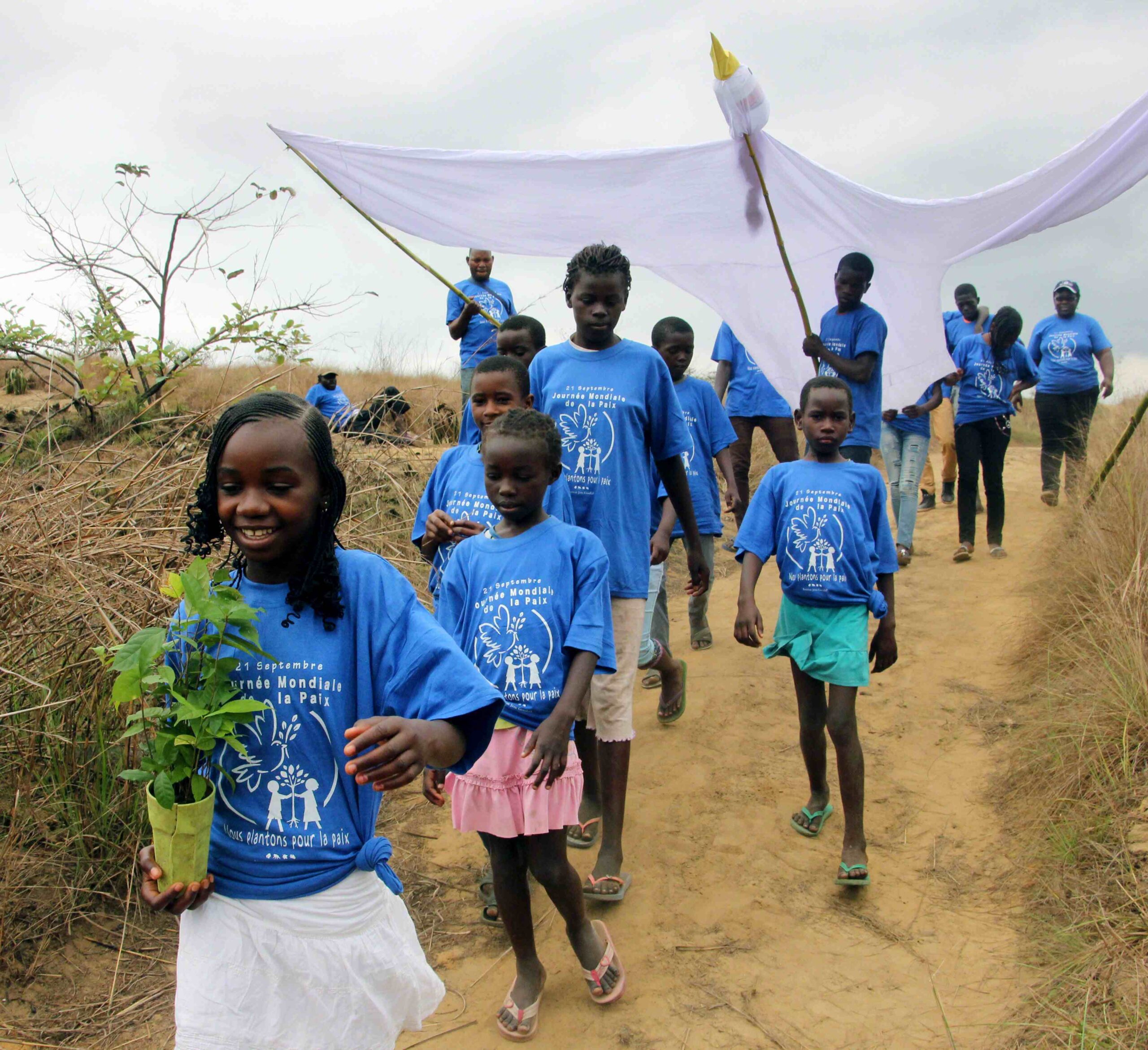 A group of about fifteen children with dark skin and black hair in matching blue shirts walking down a path, with some of them carrying a large puppet of a dove.