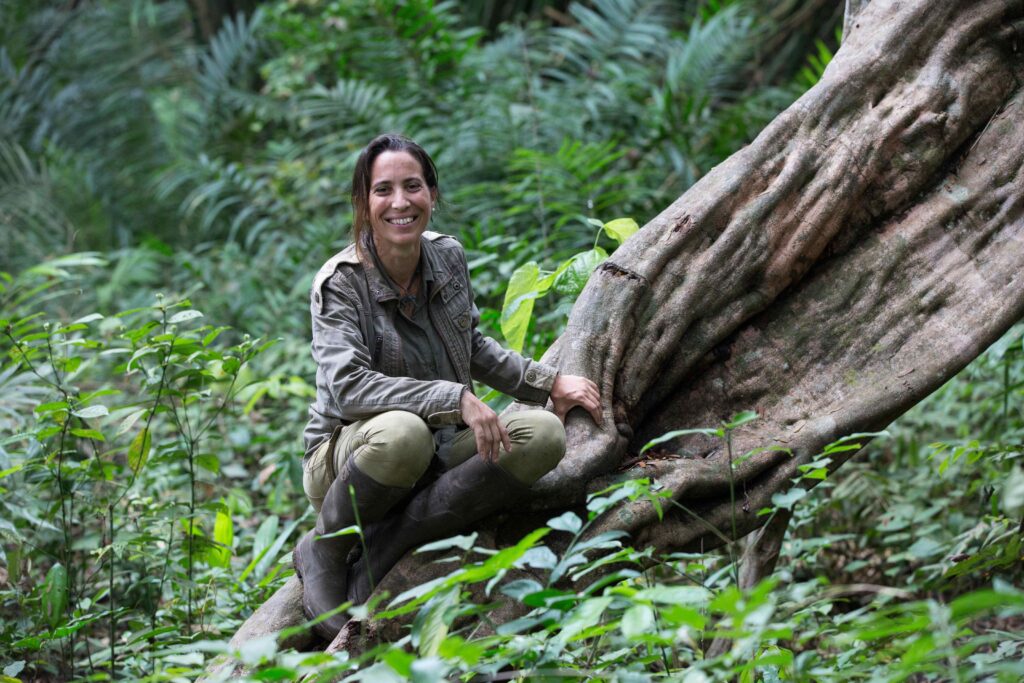 Dr. Rebeca Atencia smiling, sitting on a tree trunk in a forest setting.