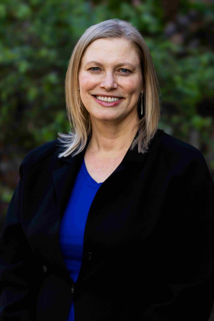 Mary Ford smiling, in a blue shirt and dark blazer in front of a leafy green backdrop.