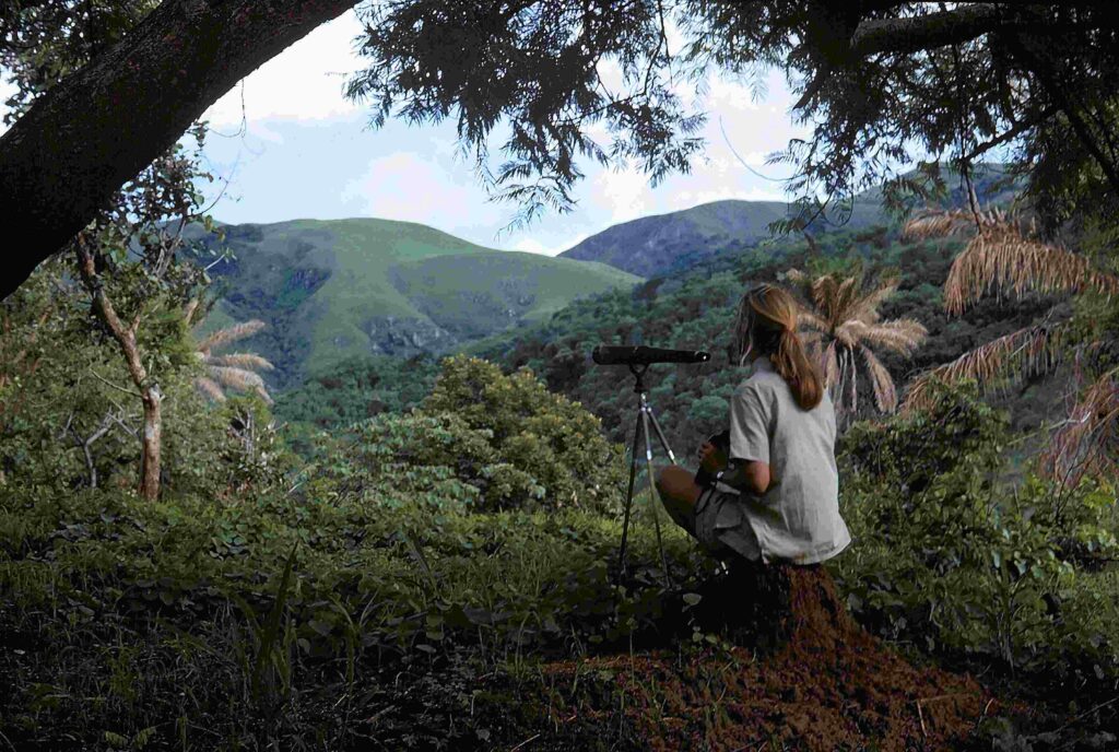 A woman with a blonde ponytail (Jane Goodall) looks through a telescope over Gombe National Park.