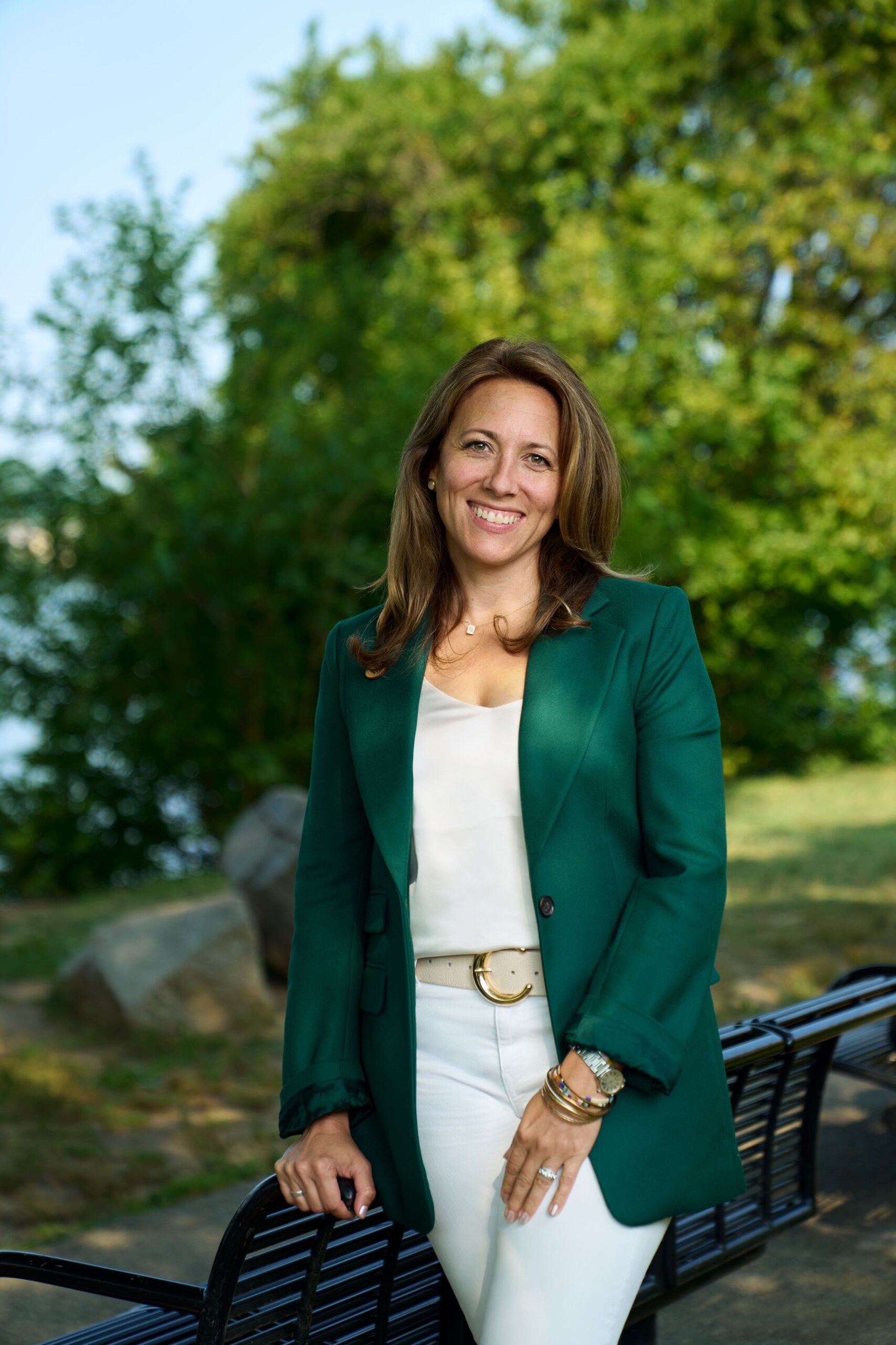 A female person (Anna Rathmann) of light complexion with dark long hair wearing a green blazer, a white shirt, and white pants in an outdoor green setting.