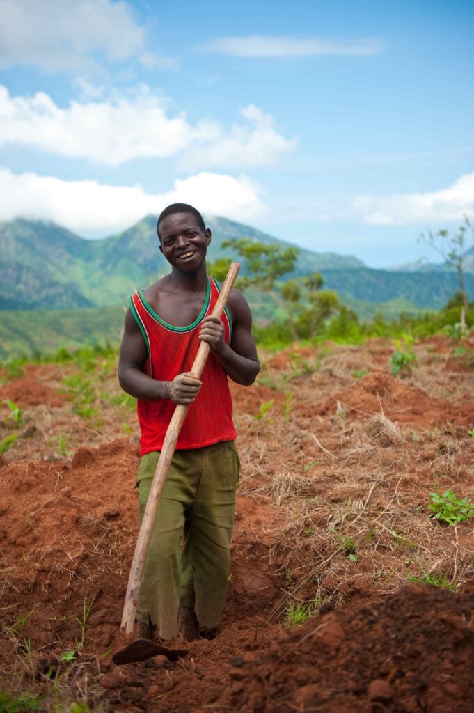 A photo of a young, masculine person with dark skin and short, curly, black hair tending to fields outside and pausing to smile at the camera.