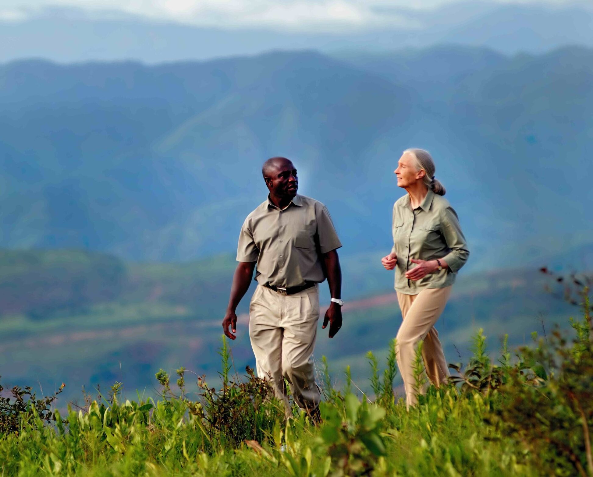 A masculine person of dark complexion and a feminine person with a grey ponytail (Jane Goodall) walk together in front of a large valley in Gombe National Park.