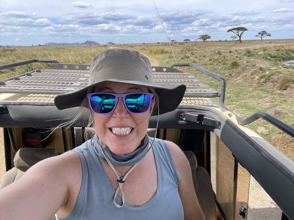 A photo of a middle-aged, feminine person with light skin and blond hair standing up in a safari vehicle. In the background is an open savannah with a few trees.