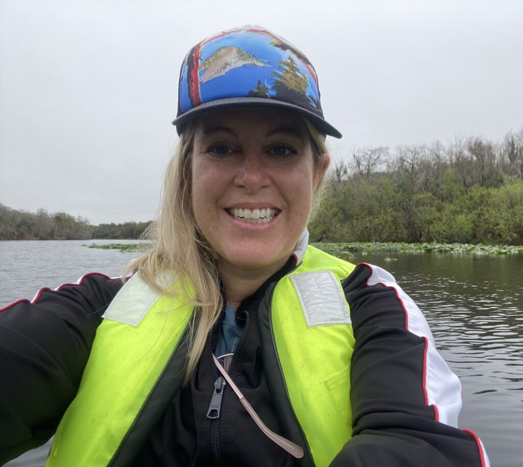 A photo of a middle-aged, feminine person with light skin and blond hair sitting in a small boat on a body of water.