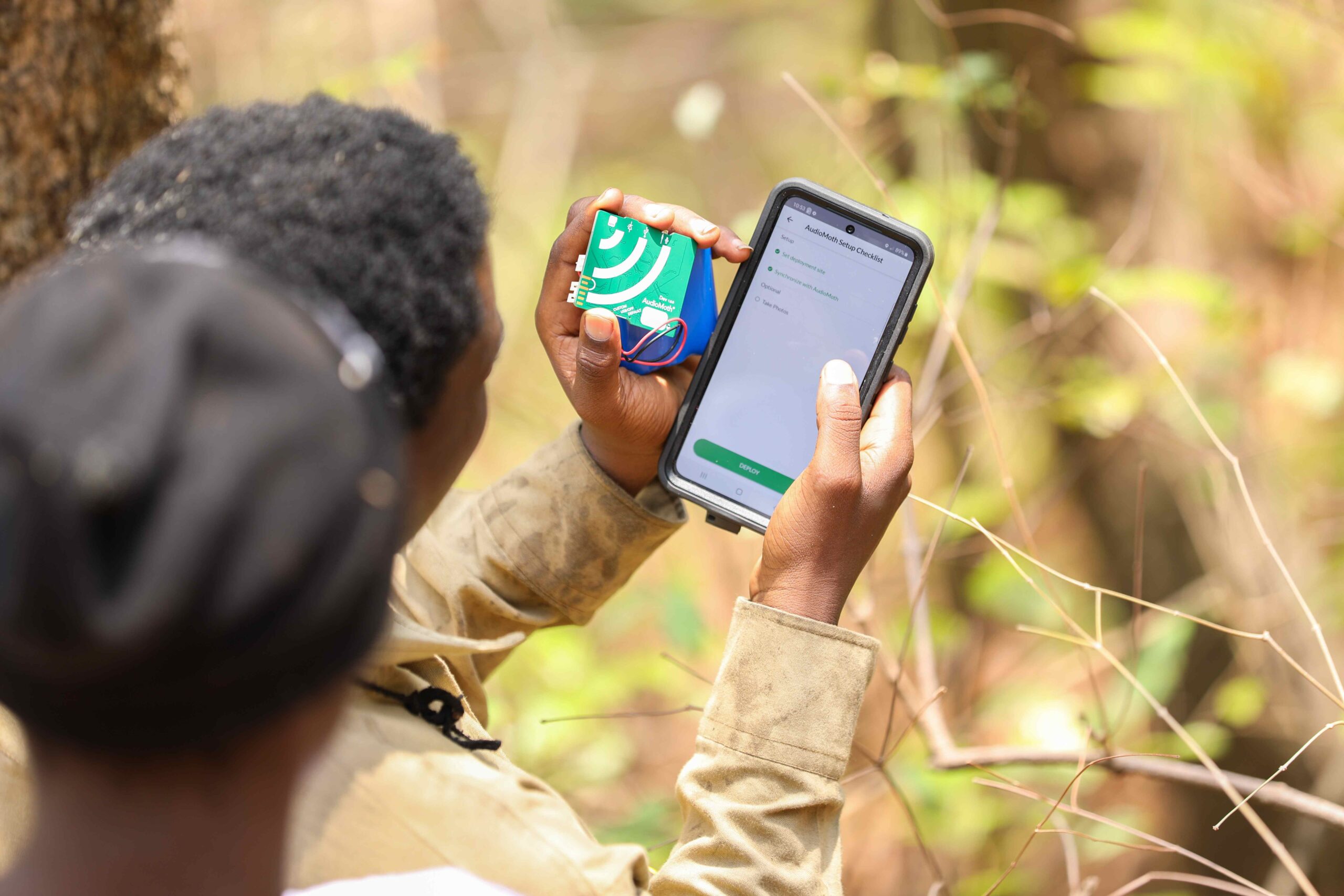 A researcher holding smart phone next to a green computer chip in an outdoor forest setting.