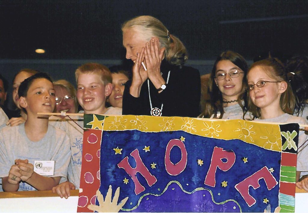 A photo of an elderly, feminine person with light skin and grey hair in a ponytail (Dr. Jane Goodall) clapping while standing in front of a group of kids and behind a handmade sign that says, "hope."