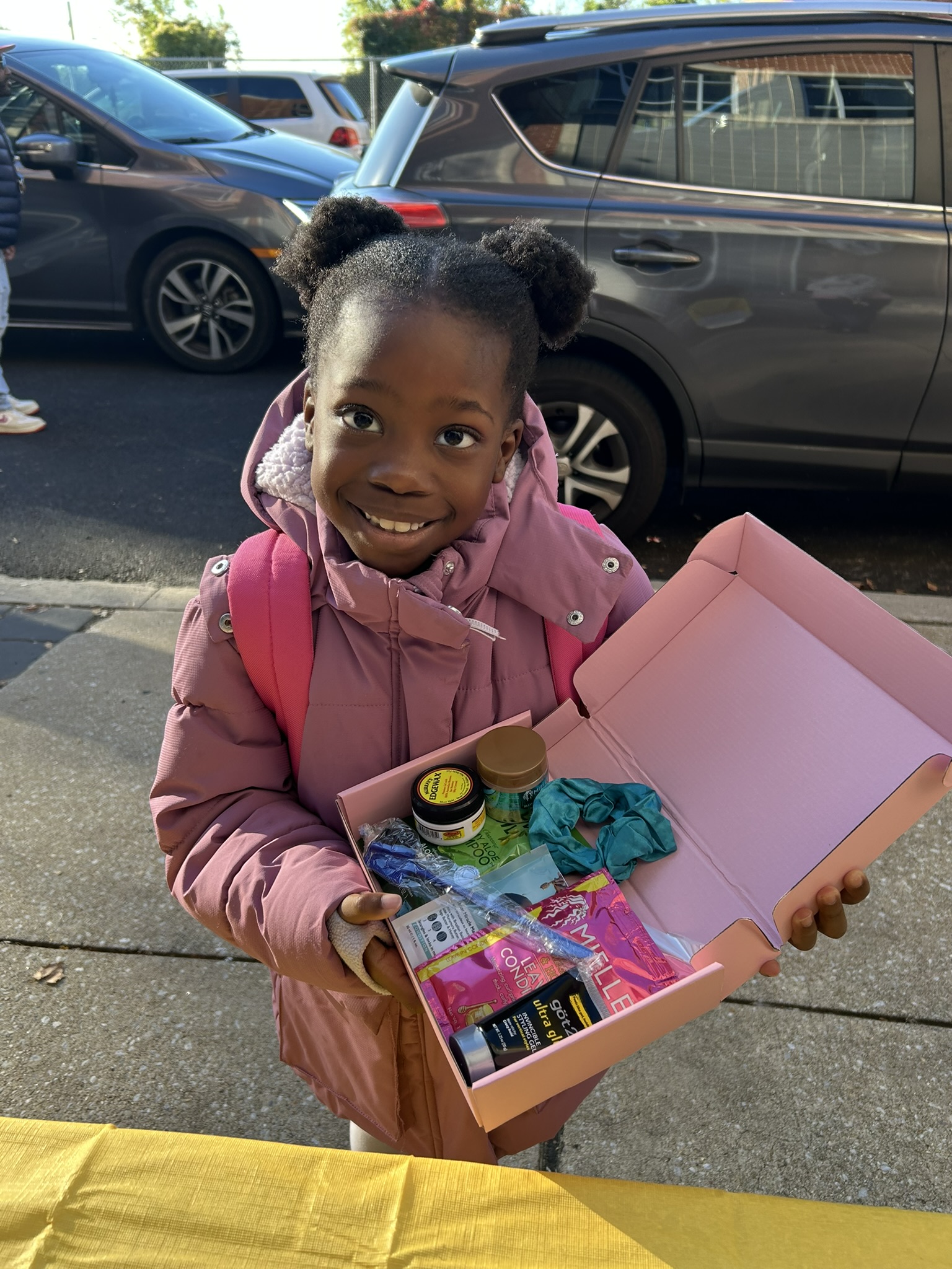 A photo of a child with dark skin and curly, black hair in two buns at the back of her head. She is holding out a box full of miniature hair products and styling tools.