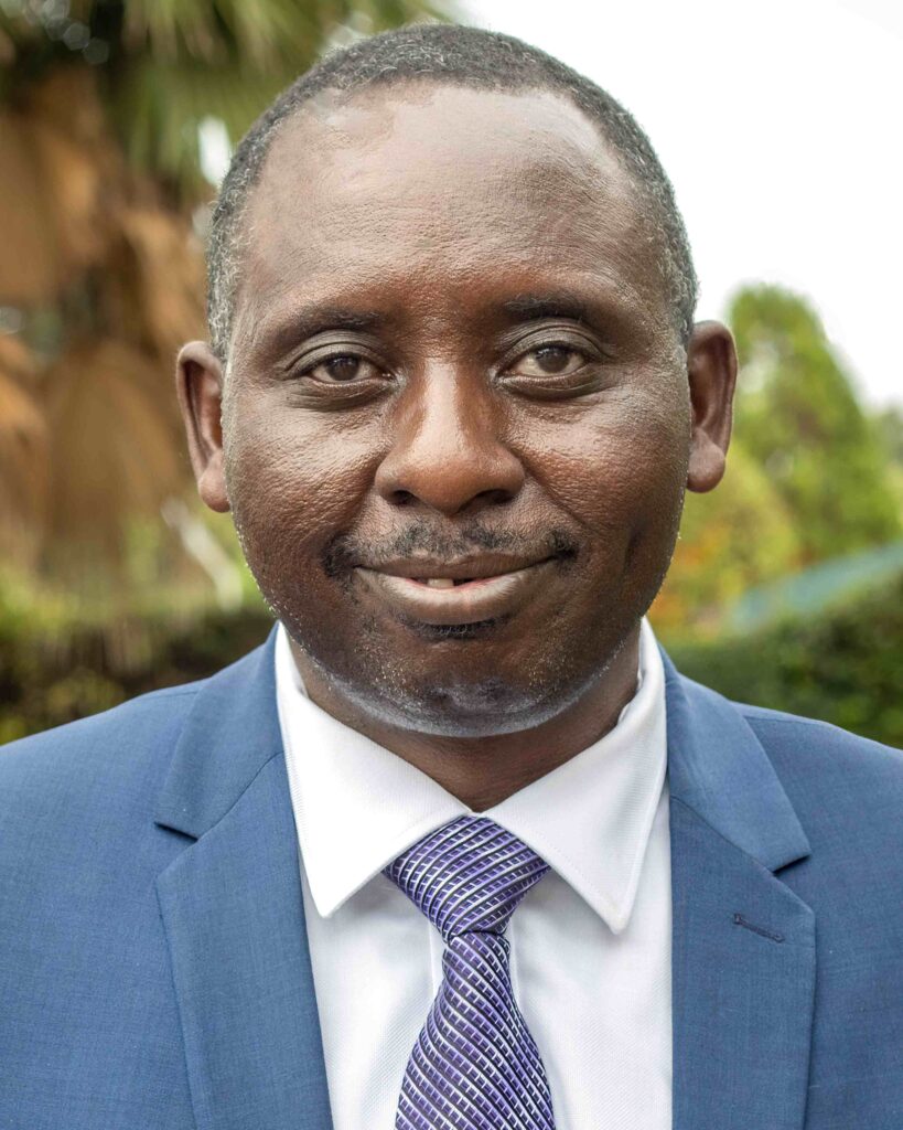 Jame Byamukama smiling, wearing a blue suit in a forest setting.