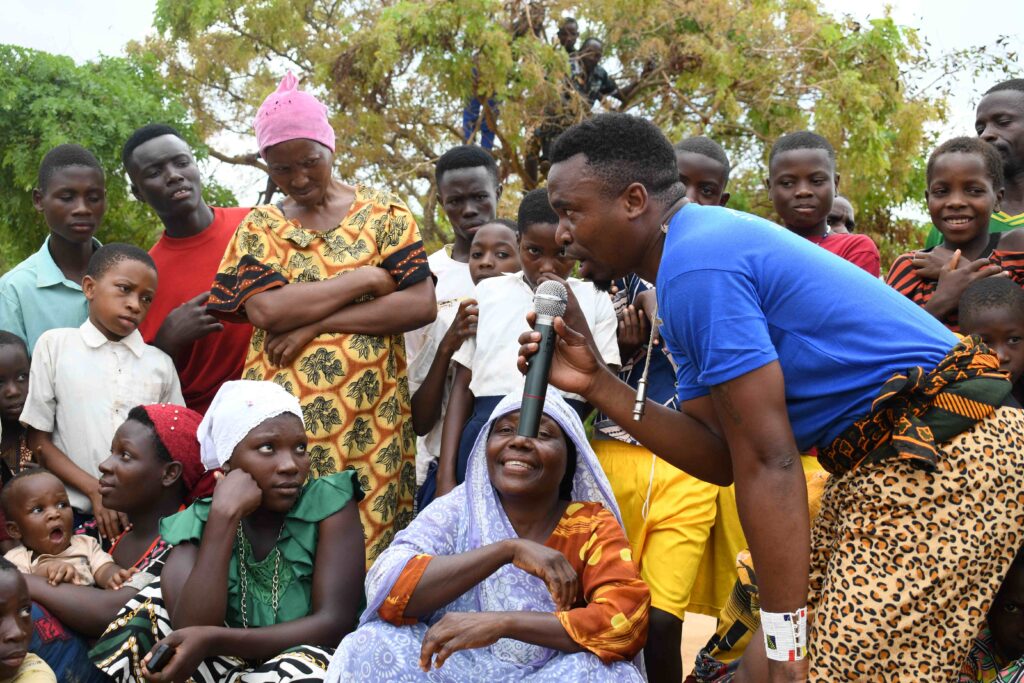 A photo of about twenty people with dark skin and colorful clothing sitting outside and listening to a middle-aged, masculine person with dark skin and short, curly, black hair speak into a microphone.