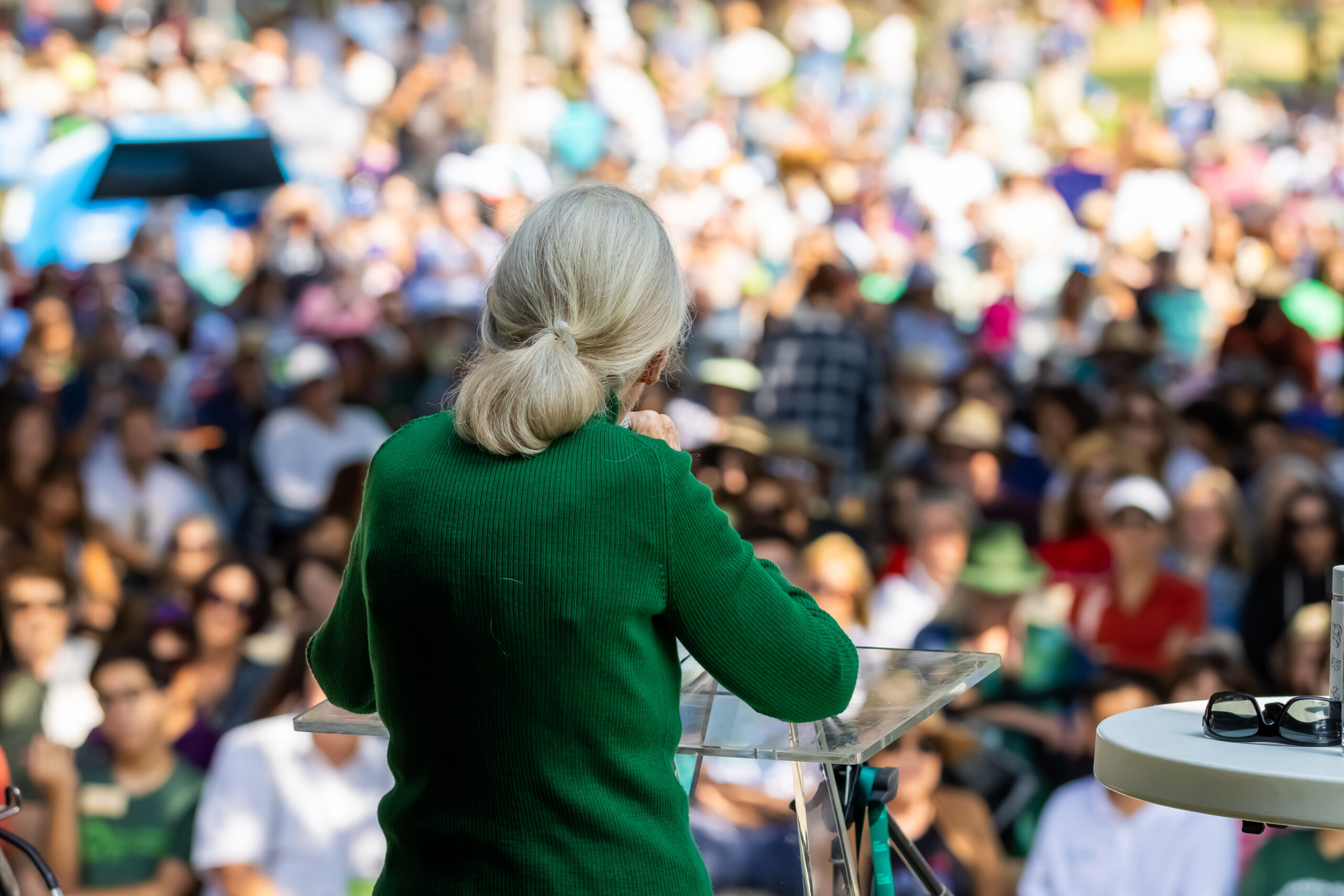 A photo of an elderly, feminine person with light skin and white hair (Dr. Jane Goodall) standing at a podium and speaking to a large crowd of people.