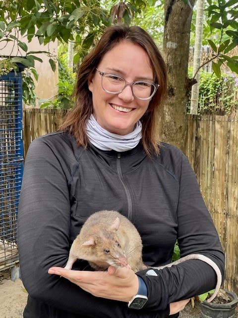 A photo of a middle-aged, feminine person with light skin and shoulder-length, brown hair standing outside and holding a large rat.