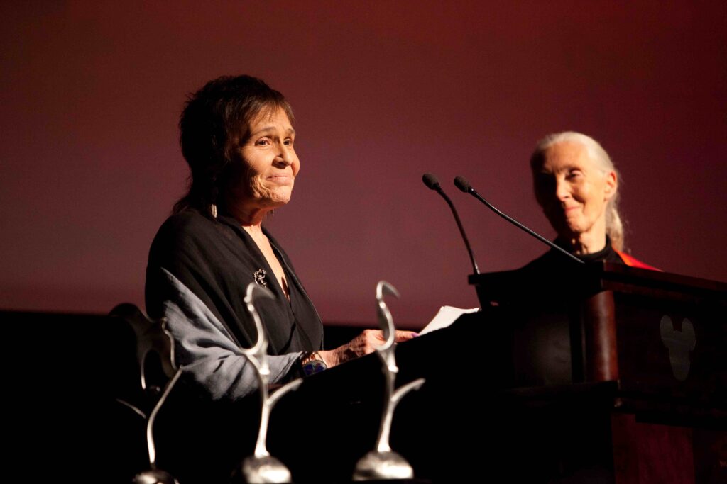 A photo of an elderly, feminine person with light skin and short, brown hair standing at a podium while an elderly, feminine person with light skin and white hair in a ponytail (Dr. Jane Goodall) listens to her.