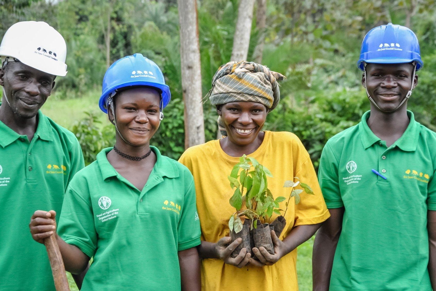 Four people stand together wearing hard hats. One holding a hoe, and one holding a plant.