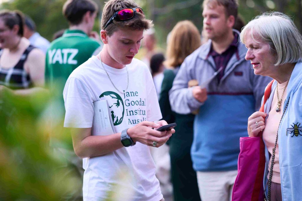 A young man with a white shirt and green text that reads "The Jane Goodall Institute Australia" shows a mature woman with grey hair his phone.
